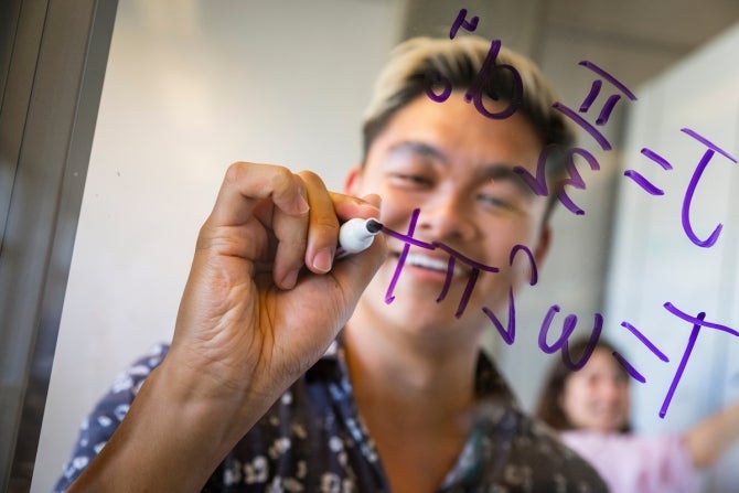 Student writing formula on window while studying with another student.